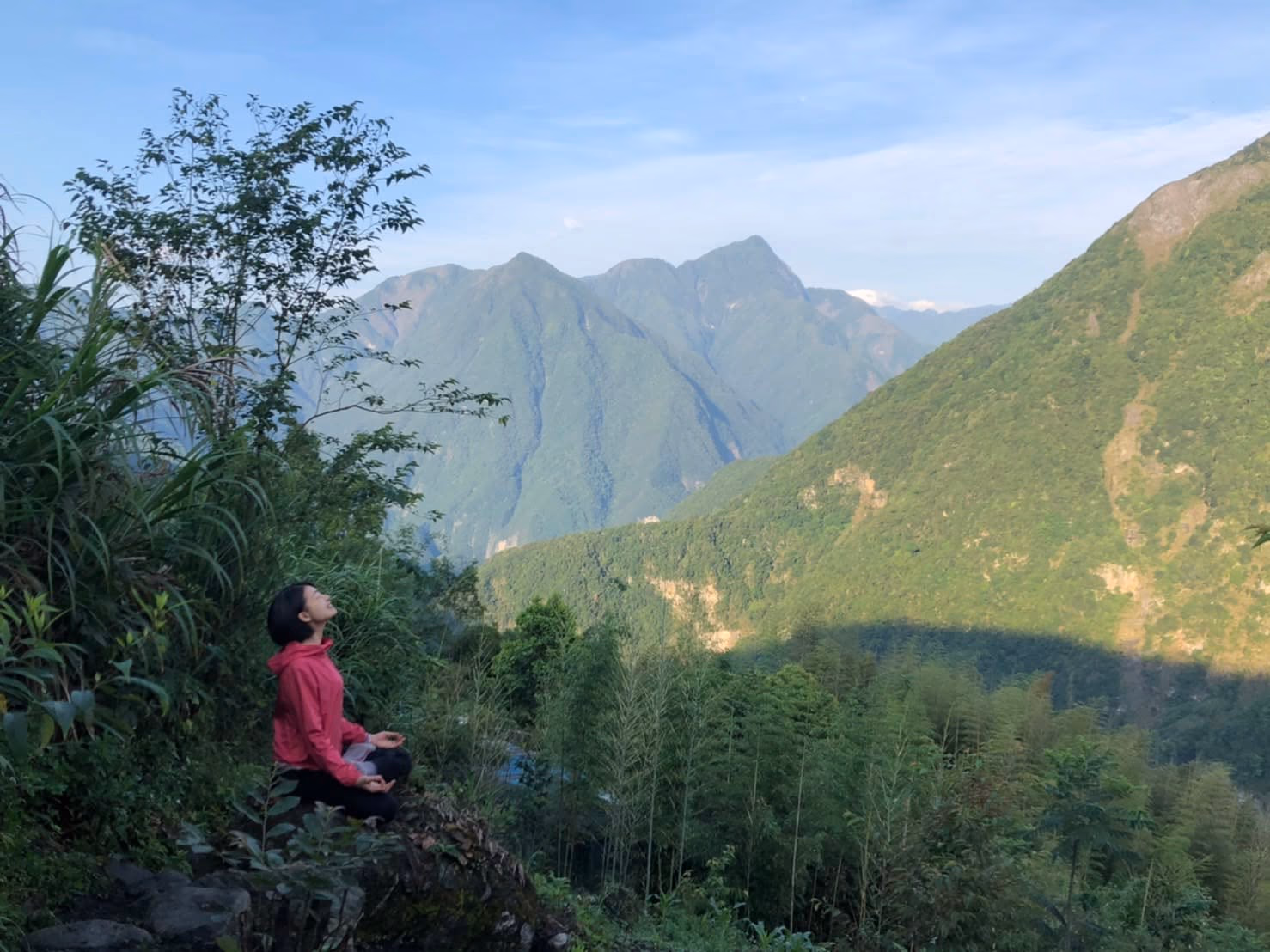 小英盤腿坐在山邊的岩石上打坐，仰著頭，背後是花蓮層層疊疊的青綠色山脈。
